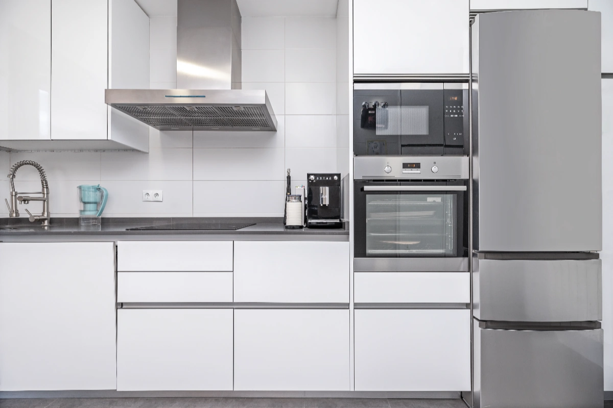 Modern white kitchen with stainless steel appliances, including a refrigerator, oven, microwave, stove, range hood, and sink with faucet. Countertop holds a coffee maker and utensil holder.