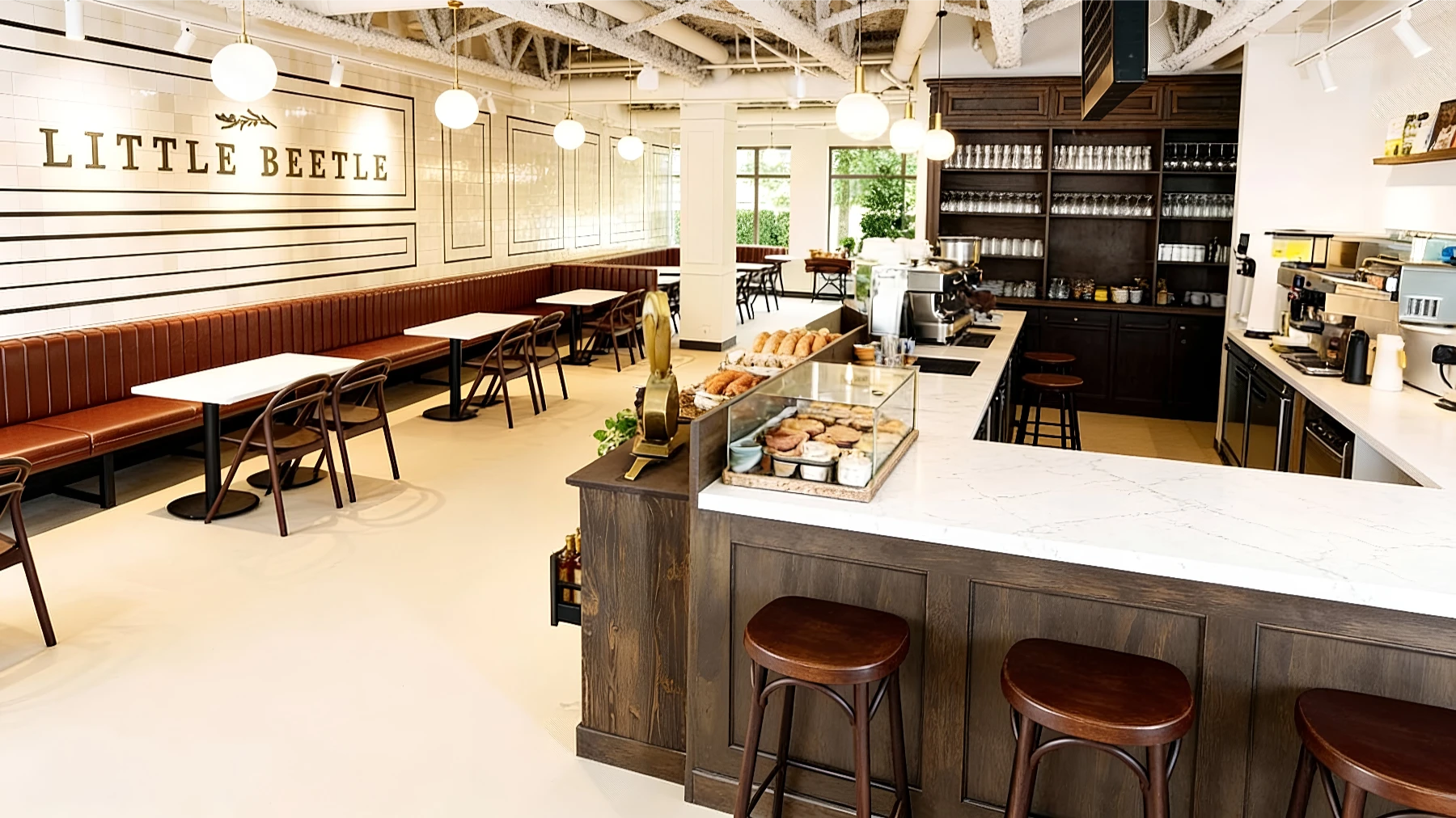 Bright, modern café interior with brown and white decor, a pastry display on the counter, wooden stools, tables, and "LITTLE BEETLE" sign on the wall.