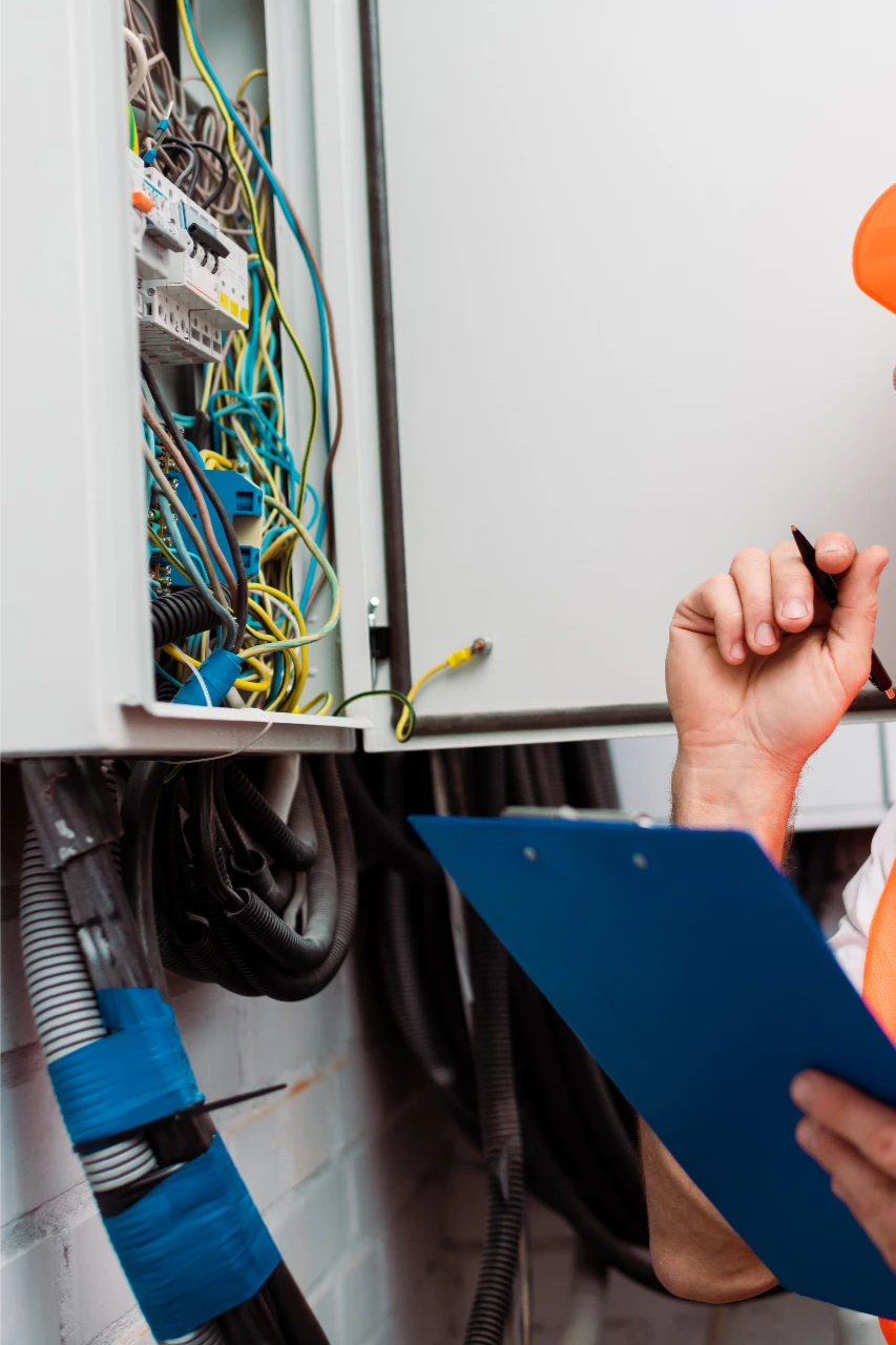 Person in a safety vest and hard hat inspects an open electrical panel with numerous wires, holding a clipboard and pen.