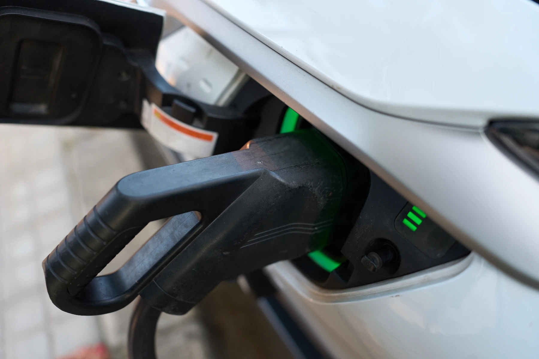 A charging connector plugged into the charge port of a white electric vehicle, with a green light indicating an active charge.
