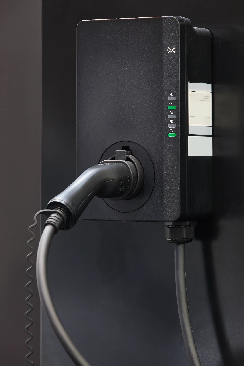A black electric vehicle charging station is mounted on a wall with a charging cable plugged in. Indicator lights are illuminated on the front panel.