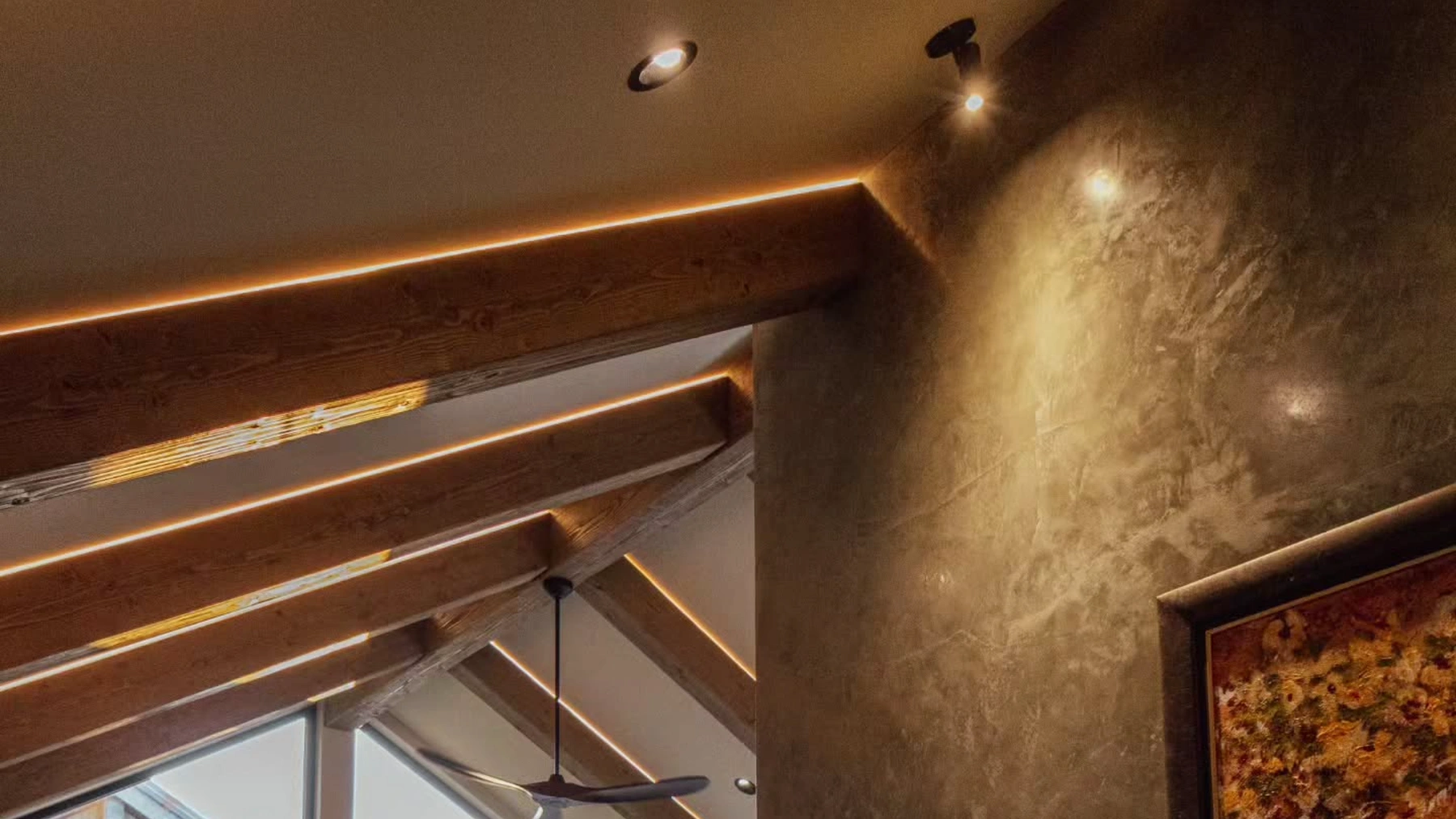 A vaulted ceiling with exposed timber beams lit by concealed LED strips, recessed pot lights, and accent lighting on a textured feature wall.