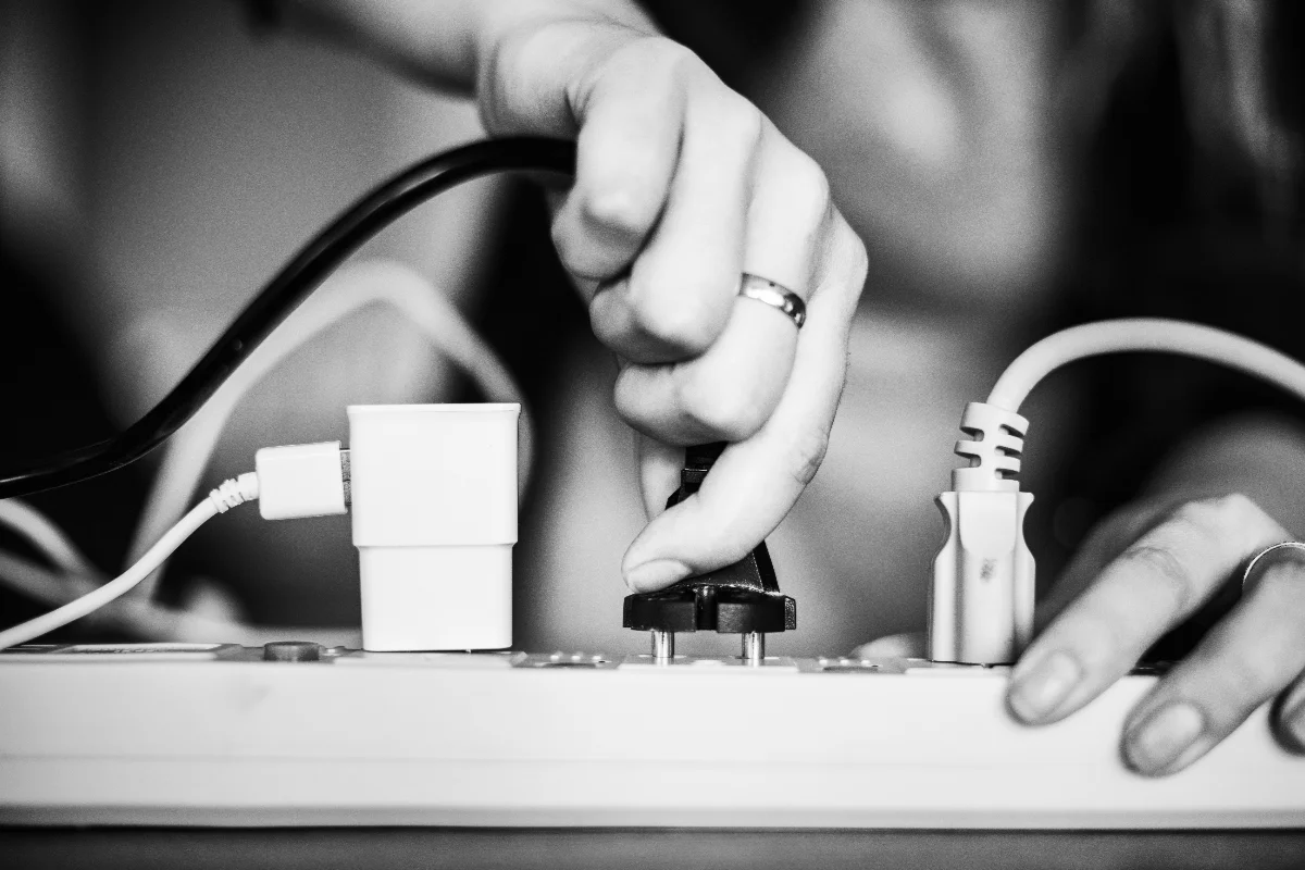 A person’s hand plugs a black electrical cord into a power strip alongside other connected plugs and adapters.