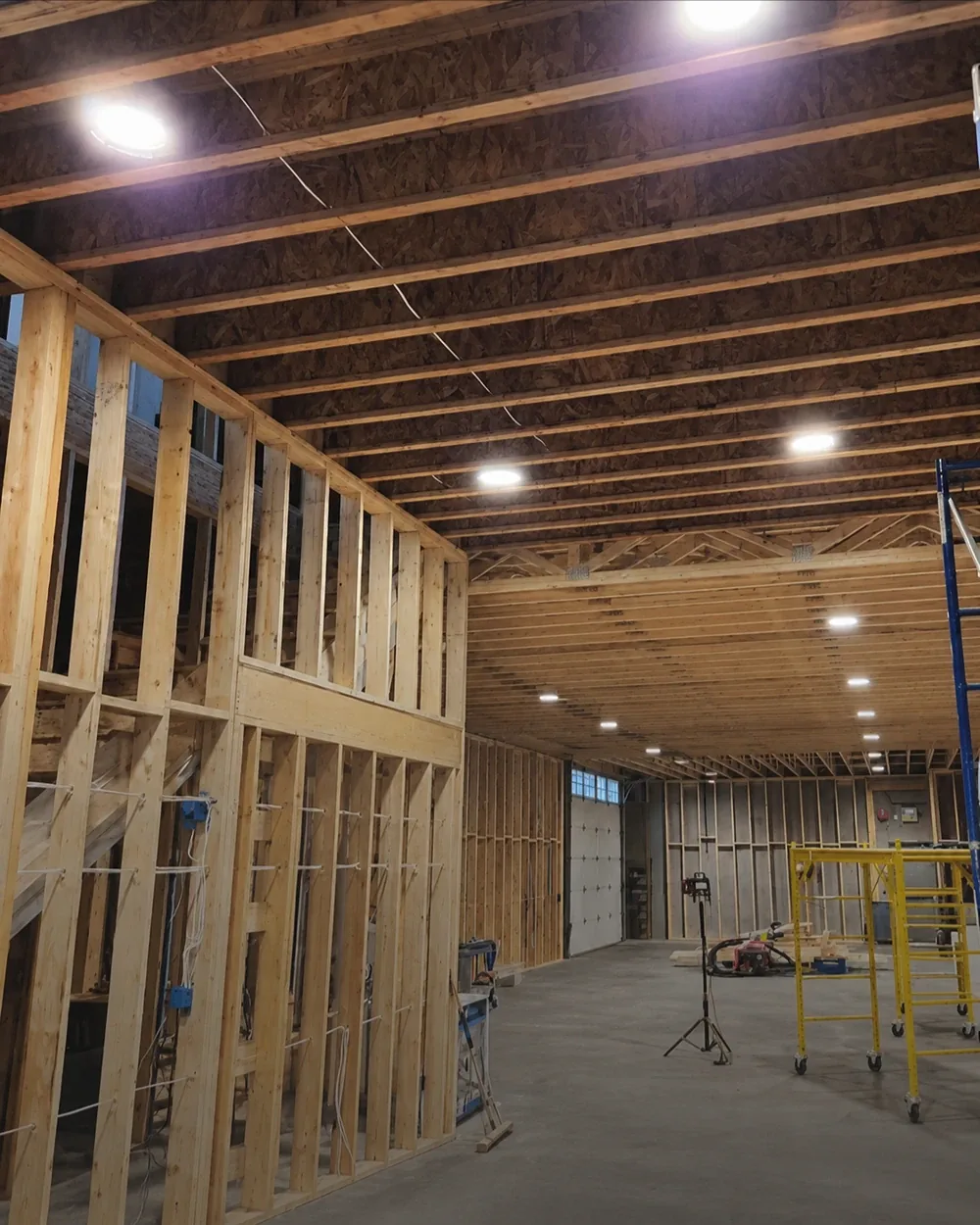 The inside of a large building still at the framing stage, lit by temporary work lights, with scaffold and a ladder set up against the open structure.