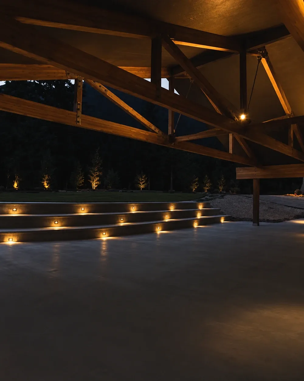 The finished pavilion at night, lit from within by the installed step and soffit lighting with the forest dark beyond it.